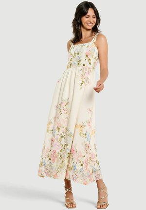 Femme souriante posant dans une robe crème sans manches, longue jusqu'aux chevilles, avec des imprimés floraux colorés et des talons dorés à brides.