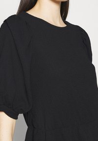 Blusa negra con tejido texturizado, escote redondeado y mangas cortas abullonadas, con un corte holgado y un sutil detalle fruncido en la cintura.