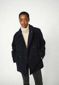 Cappotto peacoat in lana blu navy con bottoni doppi, ampie rever e tasche laterali, indossato sopra un dolcevita crema e jeans scuri.