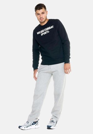Hombre de pie en pose informal, lleva una sudadera negra "WE ARE COMBAT SPORTS.", pantalones deportivos grises y zapatillas blancas sobre un fondo liso.