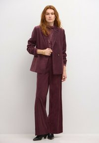 Burgundy-puku, joka on valmistettu tekstuuripinnoitteisesta kankaasta, sisältää mukautetun bleiserin, jossa on yksi nappi ja leveät housut, yhdistettynä mustiin korkokenkiin.