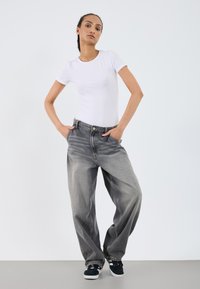 T-shirt blanc à manches courtes assorti à un jean gris délavé à coupe ample. Le modèle porte des baskets noires, les mains dans les poches, debout sur un fond neutre.