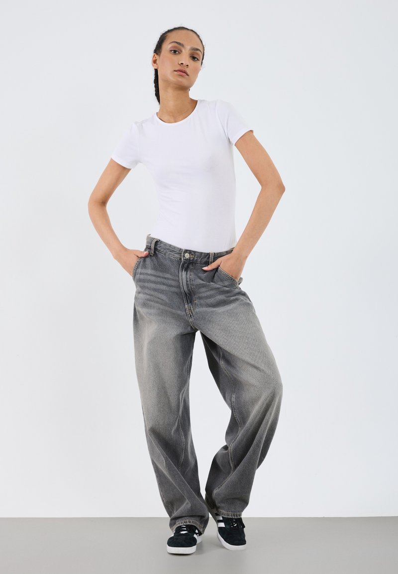 T-shirt blanc à manches courtes assorti à un jean gris délavé à coupe ample. Le modèle porte des baskets noires, les mains dans les poches, debout sur un fond neutre.