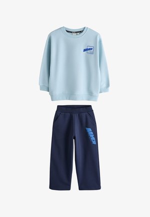 Barn med krøllet hår iført en lyseblå oversized sweatshirt med stor blå "BAKER by Ted Baker" tekst på ryggen, vendt væk.