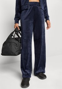 Marineblaue, samtweiche Lounge-Hosen mit weitem Bein, kombiniert mit einer schwarzen Sporttasche und schwarzen Sneakers.
