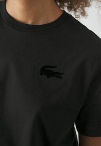 Melnas kokvilnas T-kreklu ar apaļu kakla izgriezumu. Uz kreisā krūšu daļas ir melns izšūts krokodila logo. Gluda tekstūra.