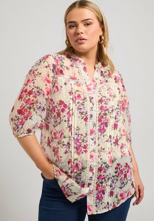 Blusa floral en tejido transparente, con flores rosas y moradas sobre un fondo crema. Incluye cierre de botones en la parte delantera y detalles de encaje.