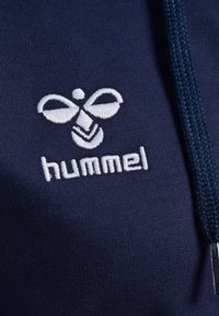 Marineblauwe stof met een opvallend wit geborduurd logo, bestaande uit twee cirkelvormige vormen en een chevronpatroon, gestikt op een gladde stoffen ondergrond.