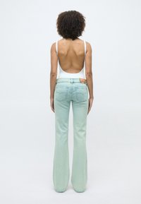 Femme aux cheveux bouclés portant un haut blanc dos nu et un jean flare bleu clair, debout de dos devant un fond uni.