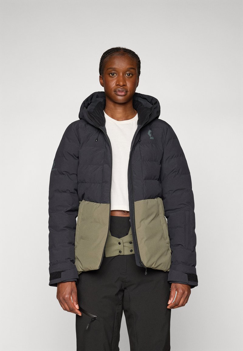 Daunenjacke mit schwarzem Oberteil und olivgrünen Hosen. Verfügt über eine Kapuze, gestepptes Muster und verstellbare Bündchen. Wird über einem weißen Hemd getragen.