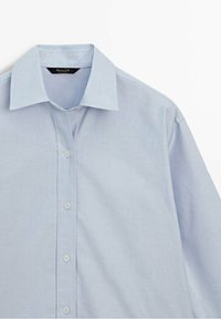 Chemise homme bleu clair à boutons, col à pointes, quatre boutons visibles et manches longues sur fond blanc.