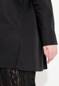 Maglietta nera con design strutturato, maniche lunghe, fessure laterali e una texture liscia. Sotto c'è un indumento in pizzo nero.