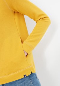 Gul texturerad långärmad topp med sidofickor, kontrasterande sömmar och en liten logotyp vid nederkanten.