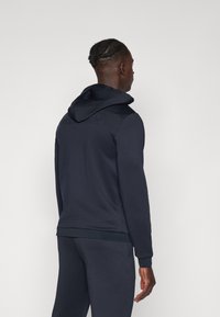 Marineblå hettegenser laget av glatt stoff, med kengurulomme, ribbestrikkede mansjetter og en brodert logo på baksiden.