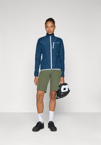 Marine vanntett jakke med hvite detaljer, grønne shorts, svarte sko og en svart hjelm med hvite accenter, stående mot en nøytral bakgrunn.