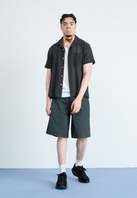 Cămașă neagră cu mânecă scurtă, cu un model texturat, purtată peste un tricou alb. Pantaloni scurți în carouri verzi și adidași negri completează ținuta.
