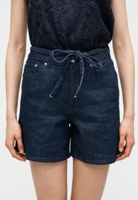 Γυναίκα που φοράει σκουρόχρωμα μπλε τζιν σορτς με δέσιμο στη μέση και μαύρο, αμάνικο, δαντελένιο τοπ, στέκεται με τα χέρια χαλαρά στα πλευρά.