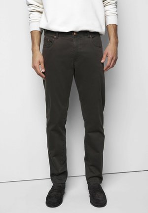 Man draagt donkerbruine rechte broek, zwarte loafers en een wit overhemd met lange mouwen, staand tegen een witte achtergrond.
