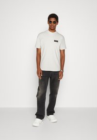 Short-sleeve, light beige t-shirt with a black logo patch on the chest, paired with dark jeans and white sneakers. Round sunglasses included.