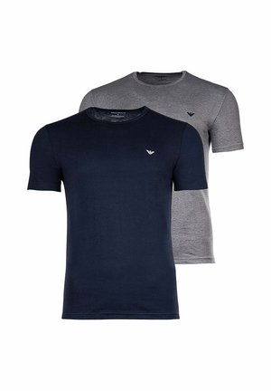 Uomo che indossa una maglietta nera aderente, pantaloni da jogging blu scuro e infradito bianche e nere, in piedi con una mano in tasca.