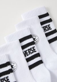 Chaussettes blanches avec des rayures noires près de la cheville, arborant un logo rond avec les inscriptions "Converse" et "Taylor" en texte noir. Texture en tricot doux.