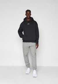 Černá mikina s kapucí a bílým logem Nike, spárovaná se světle šedými tepláky. Model stojí na jednolitém pozadí a má na sobě bílé tenisky.