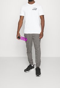 Camiseta blanca con un gráfico en negro y gris en el pecho, pantalones de chándal grises, zapatillas negras y una botella de agua morada.