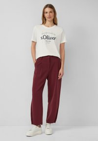 T-shirt crème avec impression graphique noire, assortie à un pantalon plissé bordeaux et des baskets blanches. Tissu lisse et coupe décontractée.