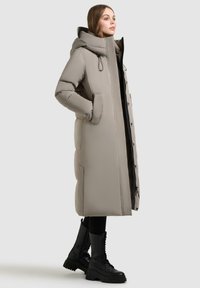 Lang, polstret frakke i lys beige med hætte, forlommer og trykknapper. Båret med sorte snørebåndsstøvler og sorte leggings.
