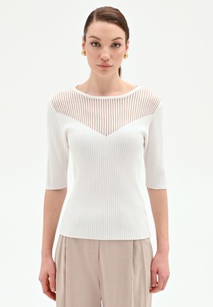 Donna con capelli castani raccolti all'indietro, che indossa orecchini dorati, top bianco a coste con scollo trasparente a righe e pantaloni beige larghi, in piedi davanti a uno sfondo bianco.