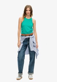 Groene mouwloze tanktop, relaxte blauwe jeans en een lichtblauw gestreept shirt om de taille geknoopt. Witte sneakers met beige accenten.