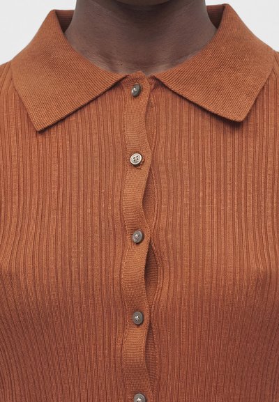 Κοντινή λήψη ενός ατόμου που φοράει ένα καφέ-σκουριάς ριμπ πλεκτό πόλο με κουμπωτό μπροστινό μέρος και διπλωμένη γιακά.