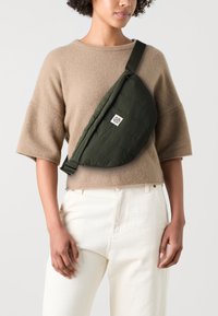 Olivengrüner gepolsterter Umhängetasche mit gesteppter Textur und einem Logo-Patch, quer über einem hellbraunen Pullover und cremefarbenen Hosen getragen.