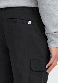 Black cargo pants with a textured finish, featuring a side pocket with a button flap and a small logo tag above a back pocket. Gray sweatshirt visible.