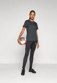 Tmavosivé tričko so zeleným logom, čierne priliehavé nohavice s textúrovanými detailmi, drží vzorovanú futbalovú loptu a má na sebe čierne futbalové topánky.