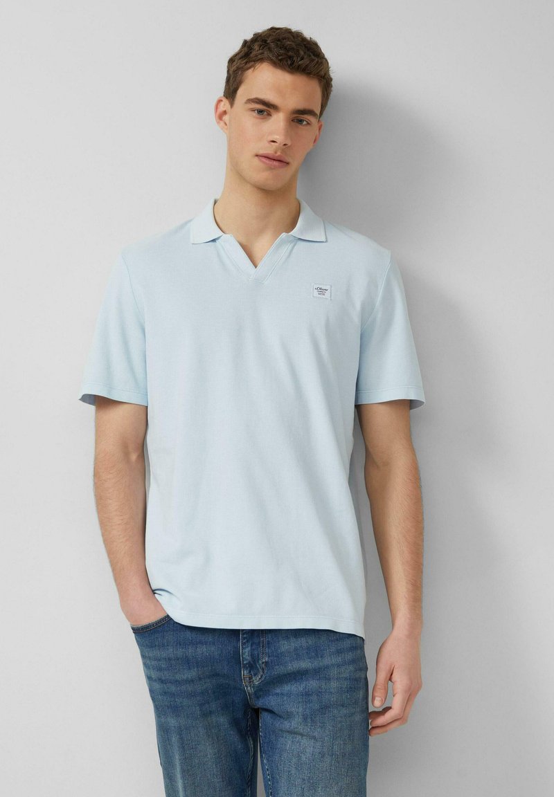 Jeune homme aux cheveux bruns courts portant un polo à manches courtes bleu clair et un jean bleu, debout contre un mur gris uni.