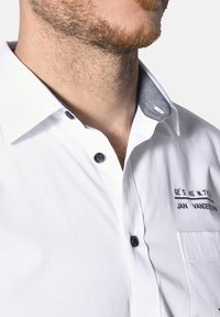 Weißes Hemd mit grauem Innenkragen. Verfügt über schwarze Knöpfe und bedruckten Text auf der Brust. Glatte, leichte Stofftextur.