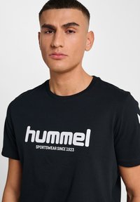Schwarzes Baumwoll-T-Shirt mit großem weißen "hummel"-Logo und dem Text "SPORTSWEAR SINCE 1923" auf der Vorderseite, Rundhalsausschnitt und kurzen Ärmeln.