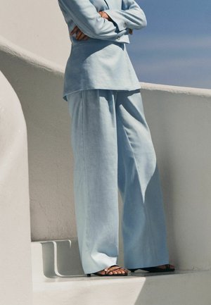 Person in light blue suit with wide-leg pants and crossed arms, standing on white steps against blue sky.
