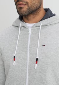 Hoodie gris clair avec une fermeture éclair à l'avant, cordons de serrage blancs avec des accents rouges et noirs, et un petit logo de marque sur la poitrine.