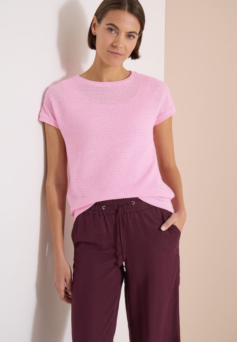 Femme portant un haut en tricot rose à manches courtes et un pantalon ample bordeaux à cordon, debout avec une main dans la poche devant un mur bicolore.