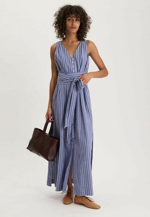 Femme portant une robe maxi sans manches à rayures bleues et blanches avec des boutons et une ceinture à la taille, tenant un sac à main marron foncé et portant des ballerines beiges.