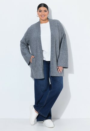 Vrouw met donker haar in een grijze open cardigan, wit overhemd, blauwe jeans met wijde pijpen en witte sneakers, staand tegen een witte achtergrond.