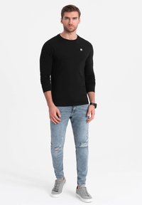 Schwarzes Langarmhemd, Slim-Fit-Design, getragen mit hellen, stark verwaschenen Jeans. Graue Sneaker, minimales Logodesign auf dem Hemd.