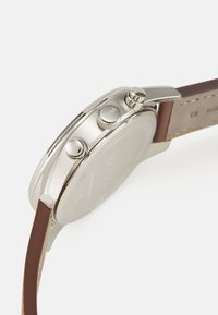 Orologio argento con cassa in acciaio inossidabile, due pulsanti e cinturino in pelle marrone. Dettaglio visibile del marchio sul lato della cassa.