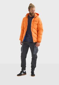 Chaqueta acolchada naranja con capucha, que presenta un cierre azul. Combinada con pantalones cargo oscuros y zapatillas negras con acentos blancos.