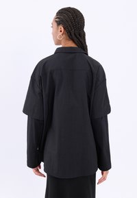 Camisa negra de gran tamaño con una textura fluida, mangas cortas y un collar clásico. La parte trasera presenta un dobladillo redondeado y un diseño minimalista.