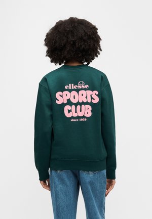 Osoba o kręconych włosach nosi ciemnozieloną bluzę z różowym napisem „ellesse SPORTS CLUB since 1959”, w połączeniu z niebieskimi dżinsami, widziana od tyłu.