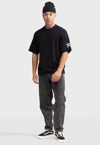 Czarna, oversize'owa koszulka z białym haftem "Rebel", w połączeniu z szarymi dżinsami i czarnymi trampkami z białą podeszwą. Na głowie czapka beanie.