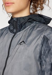 Giacca grigia leggera con finitura lucida, caratterizzata da un colletto alla coreana, cerniera frontale e un piccolo logo nero sul petto.